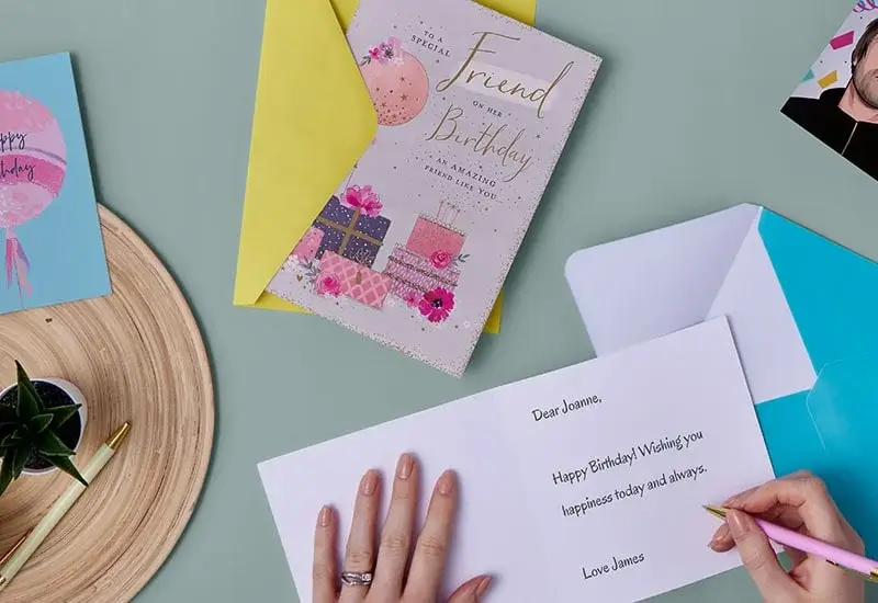 A person writing in a birthday card on a light green table, with other cards, an envelope, and a small potted plant nearby.